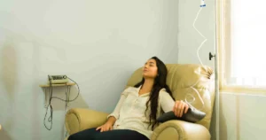 A young woman with dark hair receives IV Therapy in Meridian, Idaho, while resting in a beige chair in a light, neutral room.